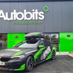 A branded black and green BMW sedan with “Apache Industrial Workwear” graphics and a rooftop cargo box is parked in front of a modern retail building. The storefront features bold signage reading “Autobits motorstore.com” in large white and green letters on a dark grey façade with bright green accents around the entrance. Clear glass windows and a landscaped parking area are visible.