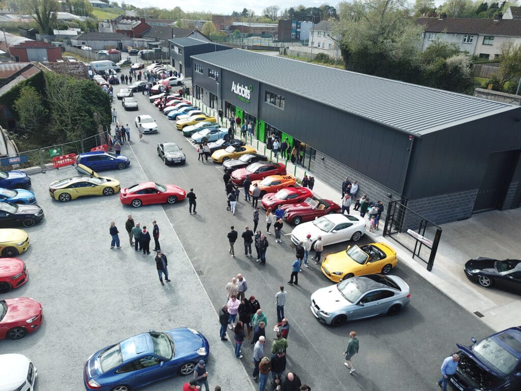 Aerial view of a car show with people walking among rows of colorful sports cars parked outside a large, modern building labeled Autobits.
