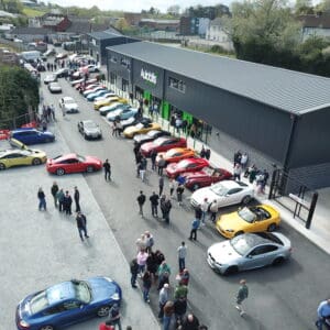 Aerial view of a car show with people walking among rows of colorful sports cars parked outside a large, modern building labeled Autobits.