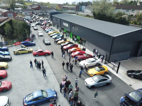 Aerial view of a car show with people walking among rows of colorful sports cars parked outside a large, modern building labeled Autobits.