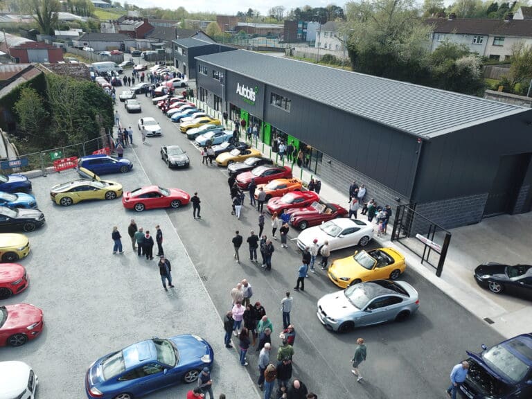 Aerial view of a car show with people walking among rows of colorful sports cars parked outside a large, modern building labeled Autobits.