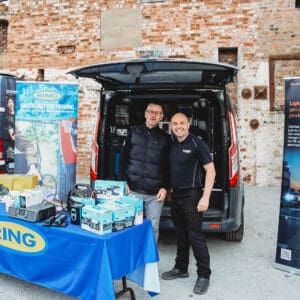 Two men standing and smiling in front of a van displaying automotive products and banners for Ring and OSRAM at an outdoor trade event.