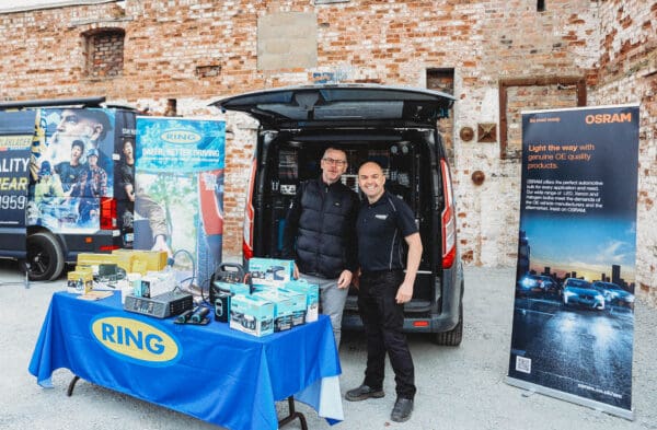 Two men standing and smiling in front of a van displaying automotive products and banners for Ring and OSRAM at an outdoor trade event.