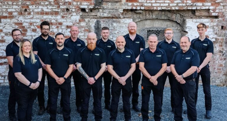 A group of 14 people standing in front of a rustic brick wall, all wearing matching black polo shirts with the "Autobits" logo. The group includes both men and women, standing in two rows, smiling and posing for a team photo outdoors on a gravel surface.