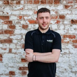 A young man (Ruari Trainor) standing with his arms crossed and smiling in front of a weathered white-and-red brick wall, wearing a black polo shirt embroidered with the ‘Autobits motorstore’ logo.
