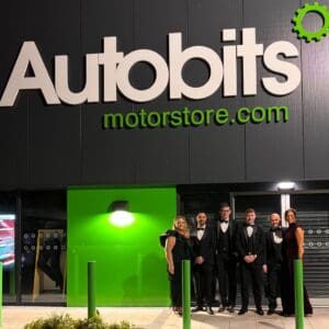 A group of six people dressed in formal evening wear standing outside an Autobits Motorstore building at night, illuminated by bright green signage and lighting