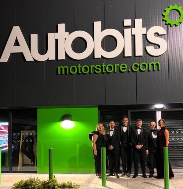 A group of six people dressed in formal evening wear standing outside an Autobits Motorstore building at night, illuminated by bright green signage and lighting